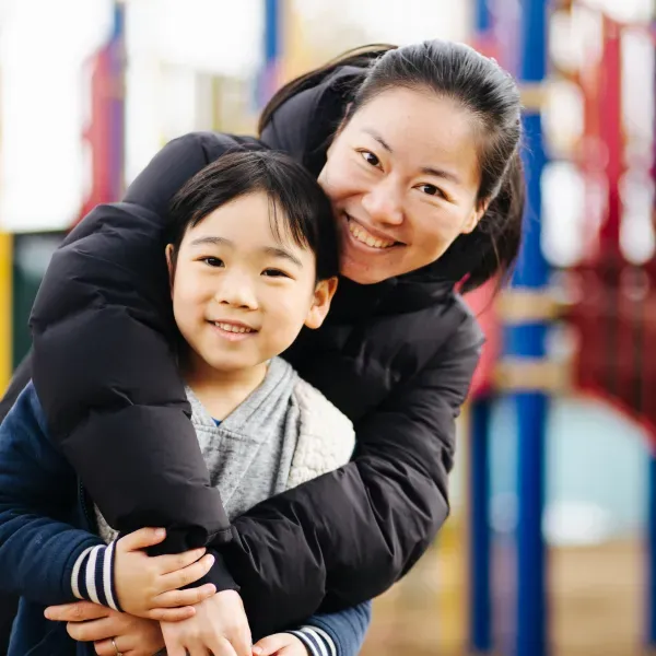 A woman, arms wrapped around her school-aged son, standing in a playground 